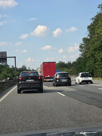 The image shows a highway scene in Rüsselsheim am Main, Germany. A black Toyota SUV is in the foreground, driving on the right lane of a multi-lane highway. The SUV's license plate reads "KUS C 638E". To the right of the SUV, a dark gray Renault Scenic is in the middle lane, with its license plate reading "MTK GI 100". Further to the right, a white Toyota Yaris is in the right lane, with its license plate reading "HR XT 8002". In the background, a large red semi-trailer truck is visible, with "KRONE" branding on its rear. The highway is bordered by trees and vegetation on the right and a concrete barrier on the left. The sky is blue with scattered white clouds, suggesting it is daytime. The traffic appears to be moving, but there are multiple vehicles, indicating it could be moderate to heavy traffic.