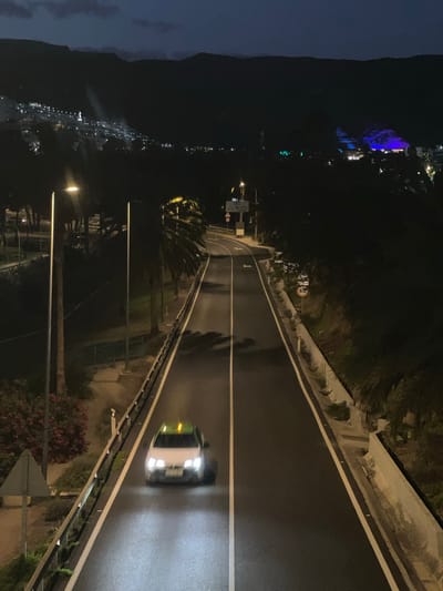 The image depicts a nighttime scene on a road in Puerto Rico, Spain. A car with illuminated headlights and tail lights is traveling on the road. Streetlights illuminate the road and surrounding areas. The road is flanked by guardrails and vegetation on both sides. In the distance, city lights are visible against a dark mountain backdrop. A "50" speed limit sign is visible near a road sign.