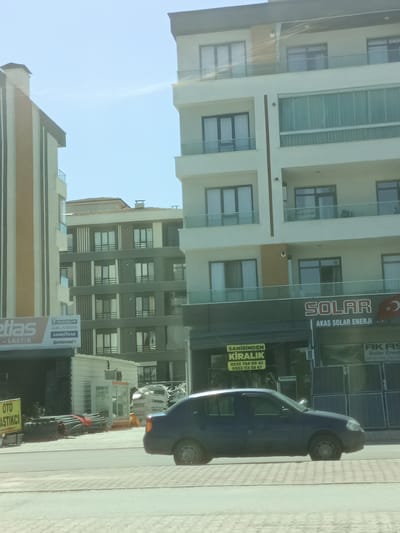 The image shows a street scene in Karatay, Turkey, likely during the daytime with clear skies. Multiple modern buildings with balconies are visible, some with distinctive architectural features like vertical striping and large windows. In the foreground, a dark blue car is driving on a paved road.
On the left side of the image, a building with "etlas" and "-LASTIK" text, along with tire brands like "MICHELIN," "ALASSA," "GOOD YEAR," and "Continental," suggests a business related to tires or automotive services. Another sign with "OTO ASTİKCİ" reinforces this idea.
In the center, a building prominently displays "SOLAR" and "AKAS SOLAR ENERJİ," indicating a solar energy company. Above this, a yellow sign reads "SAHİBİNDEN KİRALIK" with phone numbers, suggesting a rental property advertisement.
The overall scene depicts a commercial area with residential buildings, possibly a mixed-use development, showcasing a contemporary urban environment in Turkey. The sunlight suggests a bright, sunny day. There are no people clearly visible in the image, and the focus is on the buildings, signs, and the passing car.
