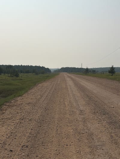 The image shows a gravel road stretching into the distance, flanked by a grassy field on the left and a line of trees on the right. The sky is overcast and hazy. In the distance, there are power lines and a wooden fence. The scene appears to be in a rural area, possibly in Myandigi, Russia, given the context provided. There are no people or animals visible in the image. The road is dry and appears to be used by vehicles, as indicated by tire tracks. The environment is natural and undeveloped.