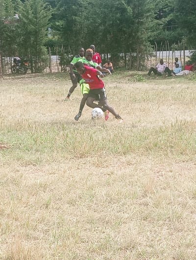 The image depicts an outdoor football match in Lwandeti ward, Kenya. In the foreground, two male individuals are actively engaged with a white football on a dry, sparsely green grass field. One player, wearing a red short-sleeved jersey with black trim, black shorts, and black athletic shoes, is in the process of dribbling the ball, leaning forward with his left foot on the ball and his right leg extended. The second player, wearing a lime green short-sleeved jersey, lime green shorts, and black athletic shoes, is positioned closely behind the first player, appearing to challenge for the ball. His hair is dyed a light blonde or yellow color.
In the mid-ground, other individuals are visible. Two men stand behind the active players; one in a green shirt and another in a red shirt. Further back, along the edge of the field, approximately six to eight individuals are seated or lounging on the ground, observing the match. They are dressed in casual attire, including shirts and trousers of various colors.
The background features a line of tall, leafy green trees, suggesting a forested or treed area. A wooden post fence runs along the perimeter of the field, visible between the spectators and the trees. To the far left, a motorcycle with a dark seat and light-colored frame is parked near the fence. The sky is bright, indicating daylight conditions.