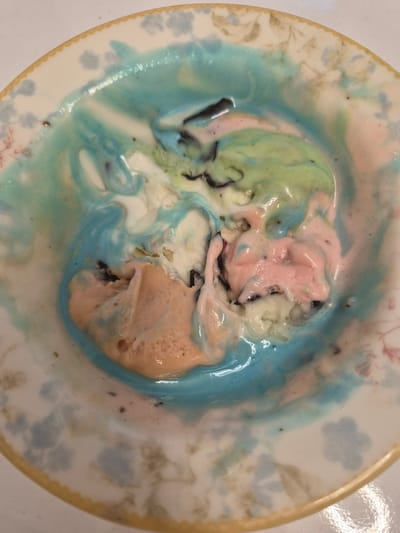 A top-down close-up shot shows a bowl of melted rainbow ice cream. The ice cream is a mix of white, light pink, light green, and light blue colors, with dark speckles throughout. The ice cream has melted and spread out, creating swirls of color in the white bowl, which has a floral pattern around its rim. The background is a white surface. The image was taken in Etimesgut, Turkey.