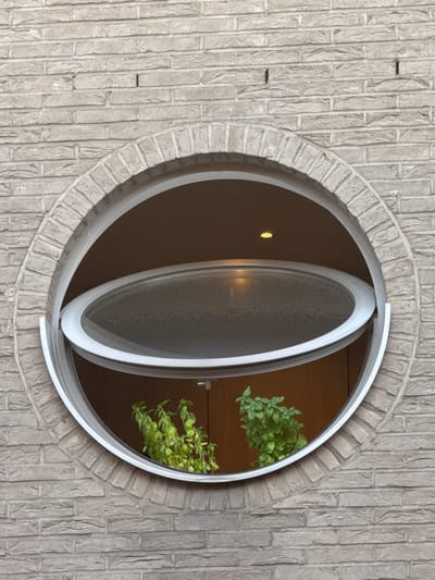 The image shows a close-up of a circular window in a brick building. The window is framed by a light-colored metal trim and has a glass pane that appears to be slightly frosted. The window is partially open, with the lower section tilted outwards. Inside the window, two potted green plants are visible. The brickwork of the building is a light grey color with visible mortar lines. The overall scene suggests an exterior architectural detail. The location is identified as Strombeek-Bever, Belgium.