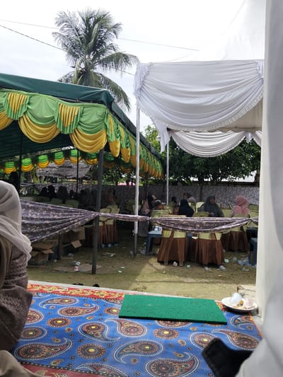 The image depicts an outdoor event, likely a gathering or ceremony, set up under two tents. The tent on the left is green and yellow with decorative draping, while the tent on the right is white with similar draping. Several people, mostly women wearing headscarves, are seated on chairs arranged around tables. The ground is covered with grass and scattered debris, including plastic bottles and cups. In the foreground, a colorful patterned rug is visible, along with a small green mat and a plate with a few items. A palm tree is prominent in the background, and the overall impression is of a casual, community event taking place in a tropical or subtropical environment. The location is identified as Cottrueng, Indonesia.