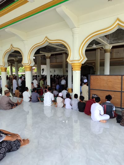 The image depicts a gathering of people, predominantly males and children, inside a mosque. The setting appears to be a covered outdoor area of the mosque, with a polished, tiled floor reflecting the light. Elaborate architectural features, including white pillars adorned with gold trim and decorative arches, suggest a mosque in Indonesia, consistent with the provided location context of Lancoklancok, Indonesia.

The individuals are seated on the floor, facing towards the interior of the mosque, indicating they are participating in a prayer or religious service. Many of the men and boys are wearing traditional Islamic attire, such as kufis (skullcaps) and long, loose-fitting garments. Some are seen in rows, while others are grouped more casually.

The activity taking place is clearly a religious gathering. The atmosphere seems peaceful and communal, with people engaged in their prayers or awaiting the start of a service. The time of day is not explicitly discernible, but the lighting suggests it could be daytime, possibly late morning or early afternoon, with no indication of overcast or specific weather conditions.

Notable details include the contrast between the brightly painted architecture and the more muted clothing of the attendees. The tiled floor is clean and reflects light, adding to the pristine appearance of the mosque's exterior. A large wooden partition is visible on the right side of the image, separating sections of the mosque or serving as a temporary barrier. There is no visible text in the image.