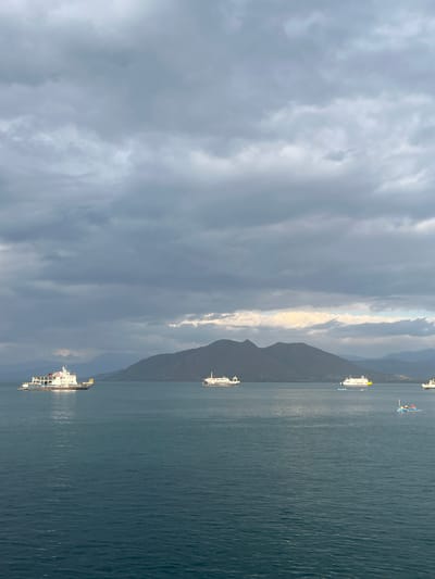 The image depicts a body of water with several vessels and a mountainous landscape in the background under a cloudy sky. The scene appears to be a coastal area, possibly near the city of Upt Tambak Sari, Indonesia, given the context.

In the foreground, the water is a deep blue-green, with subtle ripples on its surface. Several ships are visible, varying in size and type. On the left, a large cargo ship with a white superstructure and a brown hull is positioned prominently. Further to the right, two ferries or passenger ships are sailing. One is white with yellow accents, and another, slightly further back, is also predominantly white. In the middle distance, a smaller, colorful boat with a blue hull and a red canopy is also present, with at least two individuals onboard.

The background is dominated by a range of mountains, with the closest range appearing dark and rugged, possibly covered in vegetation. The sky is heavily overcast with thick gray clouds, though a patch of lighter, brighter sky is visible towards the center horizon, suggesting the presence of sunlight breaking through. The overall atmosphere is subdued, hinting at possibly inclement weather or the approach of evening. There are no discernable buildings or urban structures visible in this specific frame, but the presence of multiple vessels suggests a port or active shipping route.