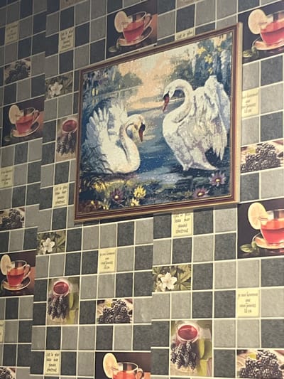 The image shows a wall with a mosaic tile pattern featuring images of tea glasses, flowers, and text. A framed picture of two swans is centrally placed on the wall. The scene does not reveal any information to suggest the location is Gorun, Bulgaria. There are no people visible.