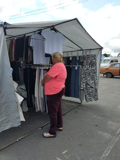 A blonde woman in a coral pink t-shirt and burgundy sweatpants stands with her back to the viewer, examining clothing at an outdoor market stall in Chudniv, Ukraine. The stall has a white tarp roof supported by metal poles and displays various garments, including t-shirts, pants, and dresses with a black and white zebra-like pattern. In the background, an orange vehicle and a white van are visible, suggesting a street or parking lot environment. The ground is paved with asphalt.