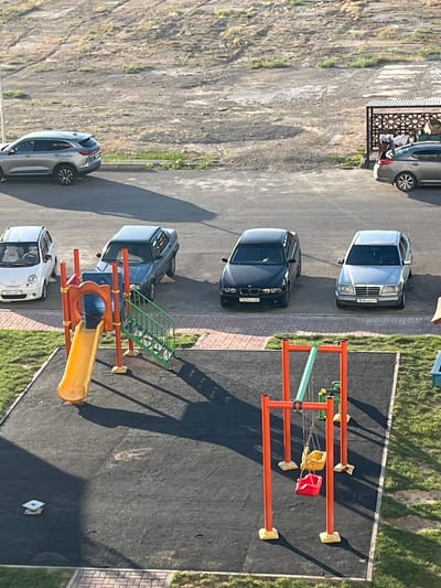 The image captures a view from above of a residential area in Türkistan qalası, Kazakhstan. In the foreground, a playground is visible with a red and yellow swing set and a yellow slide. The ground of the playground is covered with a black rubberized surface, with some patches of grass and brickwork bordering it.
Further back, a row of parked cars is lined up along a paved area. These include a silver SUV, a small white hatchback, a dark blue sedan, a black sedan, and a silver sedan. Behind the cars, a dirt and gravel embankment rises, with sparse vegetation. To the far right, a small covered structure with ornate metalwork provides shade, and two individuals are partially visible underneath it, with one of them leaning against a car. The overall scene suggests an outdoor parking lot and playground adjacent to a residential complex.