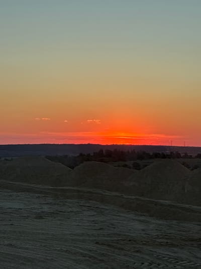 This is a landscape photograph taken at sunset in Zhirnov, Russia. The image depicts a striking sunset with vibrant orange and red hues dominating the sky. The sun is low on the horizon, casting a warm glow over the scene. To the right of the sunset, there is a dark, textured hillside that appears to be composed of rough, possibly rocky terrain. The hillside is sparsely covered with what looks like dried grass or low vegetation. The sky transitions from a soft blue or gray on the left to intense orange and red near the horizon, with some wispy clouds visible. In the distance, along the horizon line separating the sky and the land, there are faint signs of human presence, possibly structures or utility poles. The overall mood is serene and awe-inspiring, characteristic of a beautiful sunset in a natural setting. The image is oriented vertically.