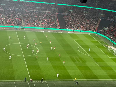 The image shows a soccer game being played on a green field inside Wembley Stadium in London, United Kingdom. The location of the stadium is at the coordinates 51.558 latitude and -0.28 longitude. The field is surrounded by a large stadium with fans in the stands, many of whom are wearing red. The teams are playing a soccer match. The players are wearing white and red jerseys. The referee is wearing a yellow and black striped jersey. There are two lines of players, one on each side of the field, and the players are running and kicking the ball. The goalkeepers are wearing blue jerseys and are standing in the goalposts. There are also two lines of officials standing on the sidelines. In the foreground, there is a small group of people standing on the sideline.