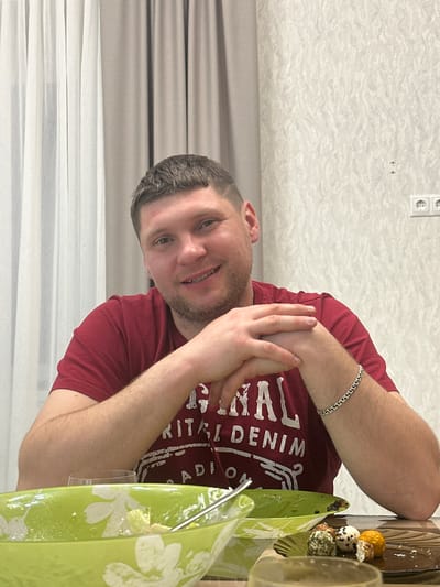 A man wearing a maroon t-shirt and a silver bracelet is seated at a table with food. The food includes decorated balls of cheese. A green serving dish with white flowers is also on the table. Behind him are light-colored curtains, and textured wallpaper is visible above the curtains. Electrical outlets are mounted on the wall. The location is presumed to be in the city of Sloboda Staraya, Russia.