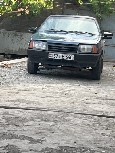 The image displays the front view of a dark blue Lada Samara (likely a VAZ-2108 or 2109 model) parked on an unpaved surface in Jrarbi, Armenia, under natural daylight. The vehicle's Armenian license plate, "AM 37 FE 640," is affixed to the front bumper. The car is oriented towards the viewer, with its front wheels visible and its lower chassis exposed.
The ground consists of coarse gravel, dirt, and fragmented concrete, with a prominent horizontal crack or seam running across the lower and mid-foreground. To the left of the car's front tire, a dried, brown leaf is visible on the ground. A thin, dark linear object, possibly a cable or hose, traverses the mid-foreground horizontally.
In the background, behind the vehicle, a light-colored, textured wall (possibly concrete or stone) is visible on the left, with some debris at its base. To the right, a darker, possibly corrugated metal or wooden structure, resembling a garage door or gate, is present. Green foliage from trees or bushes overhangs the scene at the top. No individuals or active interactions are depicted.