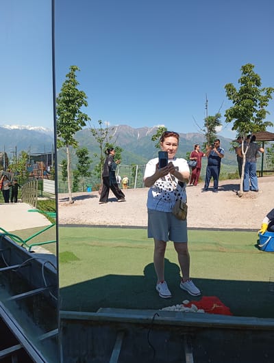 A woman in a white t-shirt with black polka dots and gray shorts is standing in front of a reflective surface, likely a window or mirror, holding a smartphone up to take a selfie. She is wearing white sneakers with red accents and has a small brown shoulder bag. The reflection shows a park or recreational area with trees, a walkway, and other people. In the background, there are snow-capped mountains under a clear blue sky. People are walking and standing around, some of them are looking at the mountain scenery. A man in a striped shirt is standing in a shaded area next to a structure resembling a gazebo. The ground is a mix of dirt, grass, and artificial turf. The lower portion of the reflection shows a metallic structure, possibly part of a fountain or water feature, with some white stones and a red cloth visible on the ground nearby. This scene appears to be located in the city of Chinarkent, Uzbekistan, based on the mountain backdrop.