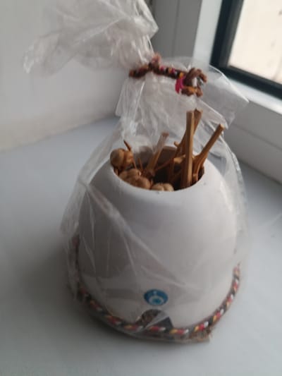 A close-up, slightly angled shot captures a white ceramic pot filled with dried twigs and small, round, brown seed-like objects. The pot is covered in clear plastic wrap, tied at the top with a colorful braided cord. The pot is decorated with a blue and white evil eye symbol. A woven cord with red, yellow, and black threads encircles the base of the pot, peeking out from under the plastic wrap. The pot rests on a white surface, possibly a windowsill, with a blurred background suggesting an indoor setting near a window. The image is taken in Arkadag City, Turkmenistan.