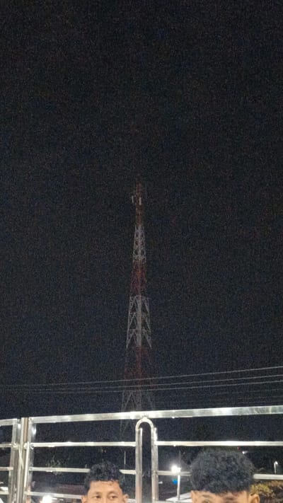 The image shows a night scene in Uteungeulinggang, Indonesia. In the background, a tall telecommunications tower stands against the dark sky, its structure illuminated with alternating red and white sections. In the foreground, a metal railing with a curved central support separates the viewer from the subjects. Two young men, partially visible from the chest up, are standing behind the railing. Their faces are turned towards the camera, and they appear to be posing for a photo. The man on the left has short, dark hair, while the man on the right has curly, dark hair. The lighting suggests it is nighttime, and the overall atmosphere is casual. There are no visible signs or text in the image.