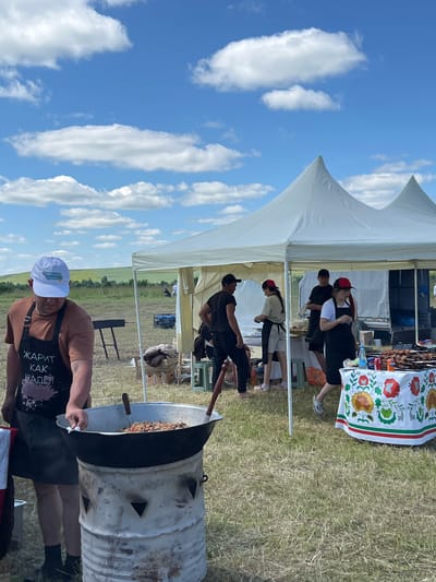 The image depicts an outdoor event, likely a food festival or market, set in a grassy field under a blue sky with scattered white clouds. In the foreground, a man wearing a white cap and a black apron with Russian text stands near a large metal pot filled with food, which is heated by a makeshift stove constructed from a metal barrel. The apron text reads "ЖАРИТ КАК НАДО!" which translates to "Fried as it should be!".
Behind the man, two white tents are set up, providing shade. Several people are visible around the tents, engaged in activities related to food preparation and service. One tent appears to be a vendor stall, with a table covered in a white tablecloth decorated with floral patterns, displaying various food items and drinks. Several individuals, including women wearing aprons and caps, are interacting near this stall. In the background, a generator and a barbecue grill are visible, along with more tents and the natural landscape of rolling hills and vegetation. The overall scene suggests a lively, community-oriented gathering in a rural or semi-rural setting. The location is specified as the city of Kalinino, Russia.