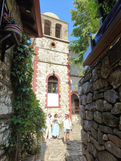 The image depicts a cobblestone alleyway in Palaios Panteleimonas, Greece. On the right, a thick, rough-hewn stone wall dominates the foreground. To the left, a stone building with red brick trim extends upwards, featuring an arched window with a white sill and small flags of Greece and another country. Further up, a bell tower with arched openings and a dome is visible against a clear blue sky. Lush green vines cascade down the stone wall on the far left. Three women are walking in the alleyway. The woman closest to the camera on the left wears a cream-colored outfit. The middle woman is dressed in a blue skirt and a striped top, carrying a yellow bag. The woman on the right wears a striped shirt, a denim skirt, sunglasses, and has pink braided hair. The sun casts shadows, indicating a bright, daytime scene.