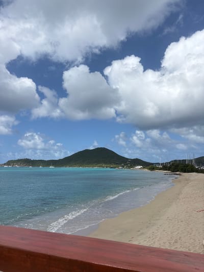 A daytime view from Spanish Town, British Virgin Islands, captures a tropical coastal scene. The foreground features a dark reddish-brown wooden railing running horizontally across the bottom of the frame. Beyond the railing, a light sandy beach extends, meeting the turquoise and blue ocean water where gentle white-foamed waves break. In the midground and background, a large, lush green, tree-covered hill dominates the center, with another smaller green landmass visible to its left. Scattered white buildings are discernible along the coastline. Several white sailboats are visible at anchor in the bay to the left, and numerous sailboat masts from a marina are evident behind a treeline on the right side of the bay. The sky above is blue, partially covered by large, distinct white cumulus clouds. No individuals are present in the image.