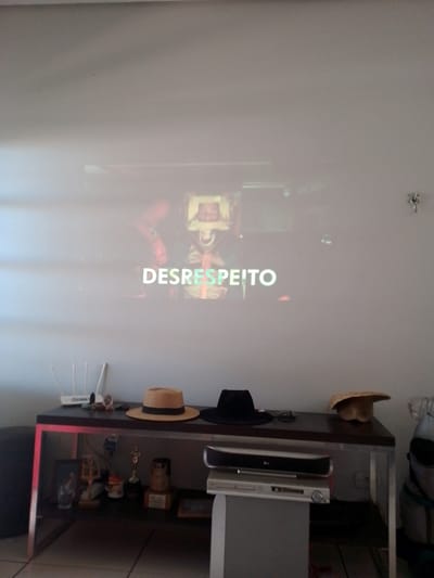 The image depicts an indoor scene, likely a living room or entertainment area, in Campo Grande, Brazil. A white or light-colored wall serves as a projection screen for a video or movie, displaying the Portuguese word "DESRESPEITO" (meaning "disrespect" or "lack of respect") in white letters. The projected image shows two individuals; one on the left is partially visible wearing an orange garment, while the person on the right is more clearly depicted from the chest up, wearing a light-colored helmet or headgear and a harness, with their mouth open, suggesting an intense or vocal action.

Below the projected image, a dark brown or black table with silver metal framing is positioned. On the table's surface, from left to right, are a white Wi-Fi router with multiple vertical antennas, a small collection of dark stones, a small framed picture, a light brown fedora-style hat with a black band, a black wide-brimmed hat, and a rustic straw hat.

The area beneath the table reveals various objects: on the far left, a dark, partially obscured object (possibly a cushion or bag), followed by a clear glass tumbler, a small wooden statuette, an unidentified white container, a silver Oscar-like trophy, another wooden object, and a small framed item.

To the right of the table, on a separate gray stand, are two stacked electronic devices: an LG-branded silver soundbar or media player on top, and a silver DVD player or similar entertainment unit below, featuring a disc tray and a control knob. An electrical outlet is visible on the wall behind these devices. Partially visible on the far right is a light-colored backpack or bag. The floor consists of light-colored tiles with visible grout lines. The overall lighting suggests an indoor setting with ambient light, possibly subdued for projection viewing.