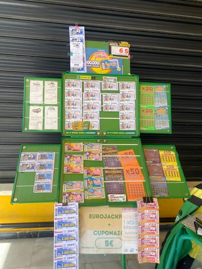 The image shows a close-up, top-down view of a lottery stand in Bel Air, Spain. The stand is filled with various lottery tickets, primarily from "ONCE" (Organización Nacional de Ciegos Españoles), a Spanish lottery and social enterprise. The tickets are displayed in green plastic trays and some are clipped together.
The subjects are primarily the lottery tickets themselves, featuring vibrant colors and different prize amounts like "GANA 500.000€," "GANA 1.000.000€," and "6.000.000€." There are also promotional banners and signs. One prominent sign reads "BOTE DE JACKPOT 100.000€ Verano," indicating a jackpot prize for a summer lottery. Another banner advertises "EURO JACKPOT" with a prize of "5€." Text on the tickets includes dates like "20 JUN 26" and "24 JUN 26," suggesting these are for upcoming draws. Some tickets are labeled "MILLONARIO," indicating a millionaire prize.
The activity taking place is the sale and display of lottery tickets, likely at an outdoor stall given the presence of a dark, corrugated metal surface in the background, possibly a storefront or stall facade. The lighting suggests it could be daytime, with natural light illuminating the brightly colored tickets. The overall impression is one of a busy point of sale for chance-based games. The text "NO TE RINDAS NUNCA ¡ILUSIÓNATE!" (Never give up, be hopeful!) and "JUEGA RESPONSABLEMENTE" (Play responsibly) appear on some tickets, reinforcing the theme of hope and responsible gambling.