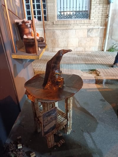 The image depicts a nocturnal street scene in El Port De Sagunt, Spain, featuring an elaborate, Harry Potter-themed display. In the immediate foreground, a large, dark brown, weathered wizard's hat with a pointed crown and wide brim is centrally positioned. This hat rests on a custom-built table constructed entirely from numerous cylindrical wine corks stacked vertically. Attached to one side of the cork table is a small, rectangular wooden sign bearing the text "Harry Potter" at the top and "PHOTOCALL" prominently in the center, with additional smaller script below.
To the left and slightly behind the hat display, an elevated platform is visible, supporting two dark, bucket-like containers. These containers are filled with white, fluffy material resembling snow or cotton, along with orange elements, suggesting a cauldron or fire effect. Golden-colored vertical rods frame this elevated section.
The background features a sidewalk composed of light-colored pavers, behind which stands a building. The building's lower section consists of light-toned stone blocks, transitioning to light brown bricks above. A window with dark, ornate metal security bars is visible on the brick wall. A white vertical downspout runs along the far right side of the building.
On the right edge of the frame, a person's lower right leg and foot, clad in blue jeans and a brown shoe, are partially visible, indicating an individual walking on the sidewalk, moving from right to left. The ground in the foreground is a dark paved surface, likely asphalt, with scattered loose corks and natural debris (leaves/twigs) near the display. The scene is illuminated by artificial light sources, casting shadows and highlighting the display elements.