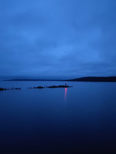 The image captures a serene, low-light waterscape, likely a lake or large bay near Medvezhyegorsk, Russia, possibly Lake Onega. The scene is set during either dusk or dawn, characterized by a dominant deep blue hue across the sky and water.

The primary subject is a small lighthouse situated on a rocky island or peninsula in the mid-ground. The lighthouse emits a vivid red light, which casts a prominent crimson reflection streak across the calm surface of the water. Surrounding the lighthouse on its small landmass are various rocks and what appear to be old, dilapidated wooden pilings or remnants of structures. To the left of the lighthouse island, more scattered wooden posts or stumps protrude from the water, suggesting submerged structures or shallow areas.

The expansive sky above is a uniform deep blue, covered with soft, indistinct clouds. Two very faint, tiny specks are barely discernible in the upper-mid section of the sky, whose nature is ambiguous (possibly distant birds or aircraft). The water itself is dark blue and largely smooth, reflecting the sky and the lighthouse's glow, indicating calm weather conditions. The distant horizon reveals a dark, undulating landmass, likely a forested shoreline or hills.

No people or direct activity are visible, lending a tranquil and somewhat isolated atmosphere to the scene. The prominent red light of the lighthouse serves as a navigational beacon in the subdued lighting. There is no visible text in the image.