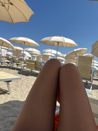 A first-person perspective shot captures sun-drenched legs resting on a beach chair, with small water droplets visible on the skin, suggesting a recent dip in the sea or a rinse. The background reveals a typical beach scene in Finale Ligure, Italy, characterized by rows of empty white umbrellas and lounge chairs neatly arranged on the sandy shore under a clear, bright blue sky. The focus is on the foreground legs, with the beach infrastructure subtly blurred, emphasizing the relaxed, vacation atmosphere.