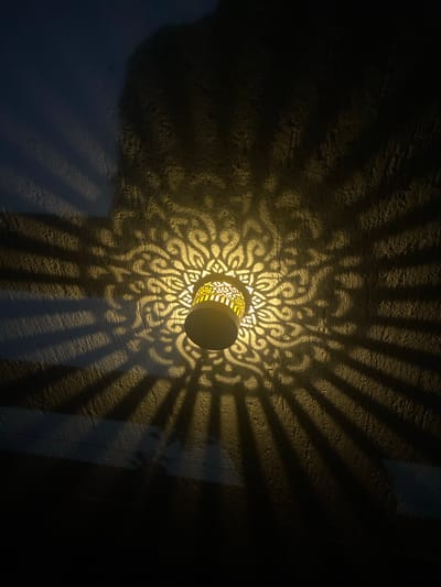 An overhead shot shows a lamp casting intricate, sunburst-like shadows on a textured, dark wall. The lamp, a cylindrical object with an ornate, perforated pattern, is positioned centrally and emits a warm yellow light. The light filters through the perforations, creating a detailed, organic pattern on the wall's surface. The shadows extend outwards from the lamp, forming radial lines and a broader, circular design that dominates the upper portion of the frame. The wall itself appears rough and uneven, adding depth and character to the projected light and shadow display. The lower part of the image fades into darkness. The location is a building in the city of Oșlobeni, Romania.
