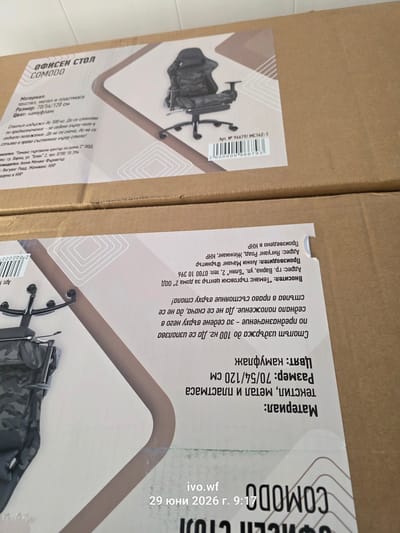 This image shows a cardboard box with text and graphics printed on it. The text is in Bulgarian and describes an office chair named "COMODO". The graphics include an illustration of the chair and a barcode. The box appears to be part of a product shipment or inventory. The bottom right corner of the image contains the text "ivo.wf 29 юни 2026 г. 9:17", which is likely a timestamp or identifier related to the photo. The box is placed on top of another cardboard box, and the overall environment suggests a storage or shipping facility. There are no people or specific locations in the city of Chal, Bulgaria, depicted in this image.