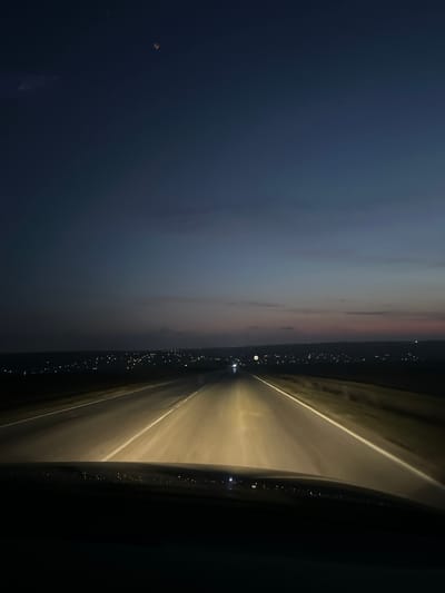 The image depicts a nighttime or late dusk scene from the interior perspective of a vehicle traveling on a paved highway. The foreground shows the dashboard or upper portion of the vehicle's interior, dark and silhouetted. The vehicle's headlights illuminate the two-lane road ahead, marked with distinct white lines that converge towards the distant horizon. Beyond the illuminated road, dark, undeveloped land borders both sides. In the mid-ground, a distant cluster of numerous, scattered white and yellow lights extends across the horizon, signifying a city. This city is identified as Kumanovo, Bulgaria. A single, distinct white light, possibly from an oncoming vehicle or a bright stationary source, is visible further down the road within the illuminated path. The sky above is a gradient of dark blue-grey, transitioning to lighter tones of purplish-pink near the horizon line. A very faint, small, orange-brown object is barely discernible in the upper left quadrant of the sky. No individuals or specific actions beyond vehicular movement are explicitly depicted.