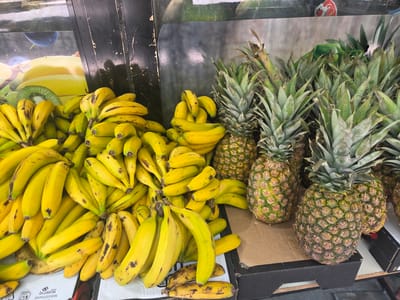 The image shows a produce stand displaying a large quantity of ripe bananas and pineapples. The fruits are arranged in piles on a white surface, with some bananas in bunches and others individually placed. The pineapples are whole and appear to be of various sizes. In the background, there is a display with images of fruits and a dark vertical structure. The lighting suggests an indoor or covered outdoor market setting. The scene is devoid of any people.