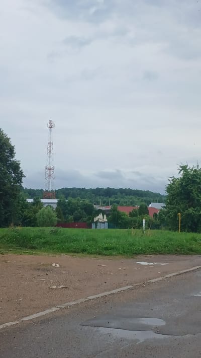 An outdoor, vertical, medium shot captures a view of a rural area under a cloudy sky. In the mid-ground, a tall, red and white lattice cell tower stands prominently. Surrounding the tower are various buildings with red roofs, interspersed with lush green trees and foliage. A dark green line of trees forms a dense backdrop against the horizon. The foreground consists of a gravel path and a portion of an asphalt road with puddles, indicating recent rain. The scene appears to be in the city of Ivakino, Russia.