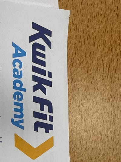 The image shows a portion of a printed document with the words "Kwik Fit Academy" written vertically in bold, dark blue capital letters. Below the text is a large, downward-pointing yellow chevron. The document is placed on a light-colored surface and is angled to the right, with a textured, light brown wooden surface visible to the right of the document. No people or interactions are visible. The location of Harderwijk, Netherlands, is not depicted in the image.