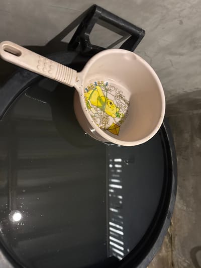 The image presents an overhead view of a beige plastic water dipper partially submerged in a dark-colored, cylindrical plastic water drum. The dipper's interior features a yellow Winnie the Pooh graphic surrounded by colored illustrations and text including "Winnie the Pooh." The dipper handle is off-white with textured grips. The water surface within the drum reflects parallel vertical light strips on the right side, suggesting illumination through blinds or a louvered opening, and a distinct circular light source reflection on the left. A black plastic handle or structural component is visible on the upper right edge of the water drum. The background consists of a light grey, textured wall. No individuals, actions, or direct interactions are present. The scene is located in Ban Mae Ping, Thailand.