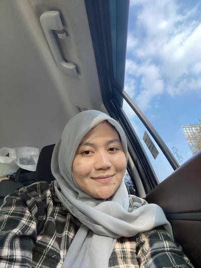 A young woman wearing a light gray hijab and a plaid shirt is seated in the passenger seat of a car. She is looking towards the camera and smiling. Sunlight streams in through the car window, illuminating her face. Outside the window, a blue sky with scattered clouds and a billboard are visible. In the background inside the car, a white shopping bag and dark luggage are present. The image appears to be taken in Mandar Sari, Indonesia, given the presence of typical Indonesian billboards and architectural styles.