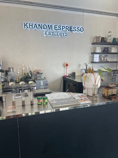The image shows the interior of a cafe, likely "Khanom Espresso EST. 2012" as indicated by the illuminated sign on the wall. The cafe appears to be located in Khanom, Thailand, given the name of the establishment. The scene is a counter area with various items for preparing and serving coffee and tea.
On the counter, from left to right, there are three glass vials containing what appears to be ground coffee or tea, labeled "Colombia," "Kenya," and "Chocolate Blueberry." Next to these are two green cylindrical containers, possibly for matcha or tea, with Chinese characters on them. Further to the right, a menu is displayed on a clear acrylic stand. Behind the counter, there are dispensers, bottles, a scale, and a cash register.
Above the counter, on shelves mounted on the wall, are various items including what looks like coffee beans in bags, a Marshall speaker, and a glass siphon coffee maker. The wall behind the counter has a textured finish. The overall environment suggests a modern, clean cafe setting. There are no people visible in the image.