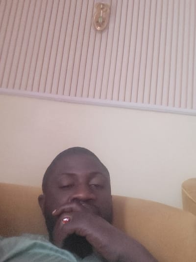 The image depicts a single male subject in an indoor environment, located in Babawa, Nigeria. The subject has a dark complexion and is shown from the chest up, in a seated or reclining position on a yellow or mustard-colored upholstered furniture item. He wears a light green short-sleeved garment. His right hand is positioned near his chin or mouth, and a ring with a prominent red stone is visible on his right ring finger. The background consists of a plain, light-colored wall. Above the wall, a white ceiling features distinct vertical linear grooves or panels. A small, ornamental, gold-toned light fixture, containing a central green illuminated element or detail, is mounted on the upper portion of the grooved ceiling.