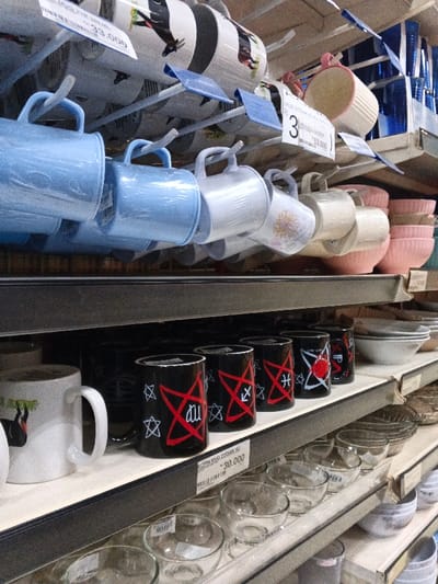 This media file captures a close-up view of shelves inside a retail store, likely a homeware section, in Sindanglaya, Indonesia. The scene is brightly lit, showcasing various kitchenware items.
The upper portion of the image features a display of mugs hanging from metal racks. On the left, several light blue mugs are visible, possibly made of plastic or ceramic, some appearing individually wrapped. To their right, white or off-white mugs with different designs, including a floral pattern, are also displayed. Further right, stacked pink bowls with a ribbed texture are arranged on a shelf. Several price tags are attached to the racks; one near the blue mugs reads "MUG-ZODIAK SG" and "33.000", while another to its right shows "33.000" and "53.000" with a "3" badge.
The lower shelf prominently displays a row of black mugs featuring distinct red and white designs. These designs include a stylized red Arabic script for "Allah" (الله) enclosed within a red pentagram-like symbol, accompanied by small white stars. Other black mugs in the row display similar red pentagram-like shapes with astrological symbols, such as Sagittarius (an arrow) and Pisces (two fish). To the left of these black mugs, a white mug with a black and white design is partially visible. In front of the black mugs, a price tag states "KOPIN MUG ZODIAK SG" and "30.000". Below this shelf, and on the next shelf down, numerous clear glass bowls of various sizes are neatly stacked, indicating a variety of glass kitchenware available. No people or activities are depicted in the image.