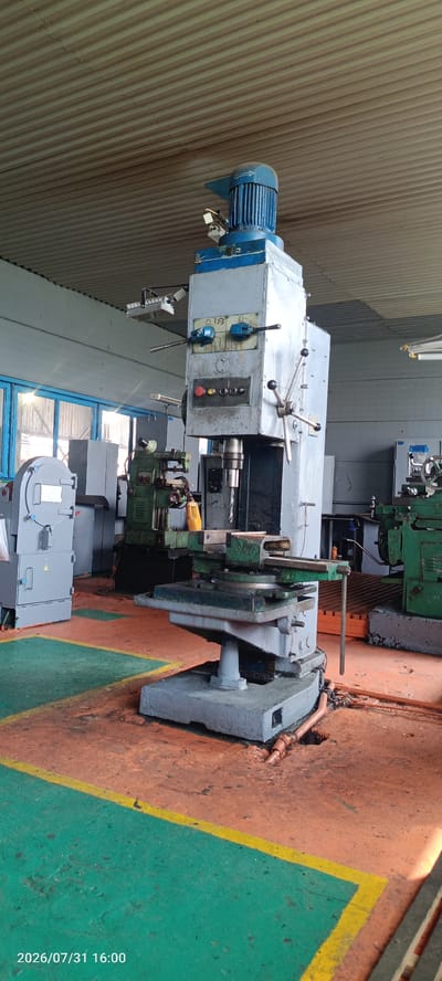 The image shows a large, grey, and blue industrial drilling machine with a blue motor at the top. The machine is in a factory or workshop environment with other machinery visible in the background, including a green industrial machine and a grey electrical cabinet. The floor is painted with orange and green sections separated by yellow lines. There are also windows with blue frames visible in the background. The scene appears to be dated 2026/07/31 at 16:00, with the timestamp overlaid on the bottom left of the image. There are no people visible in the image. The overall environment suggests a manufacturing or repair facility.