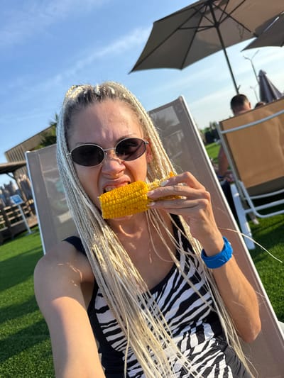A blonde woman with long, thin braids is sitting in a lounge chair outdoors. She is wearing sunglasses and a black and white zebra-print swimsuit. She is eating a piece of corn on the cob, with her mouth open and her tongue sticking out. Her left arm is bent, and she is holding the corn in her hand. She is wearing a blue bracelet on her left wrist. The background shows a grassy area, lounge chairs, umbrellas, and a clear blue sky. The location is the city of Dumino, Russia.