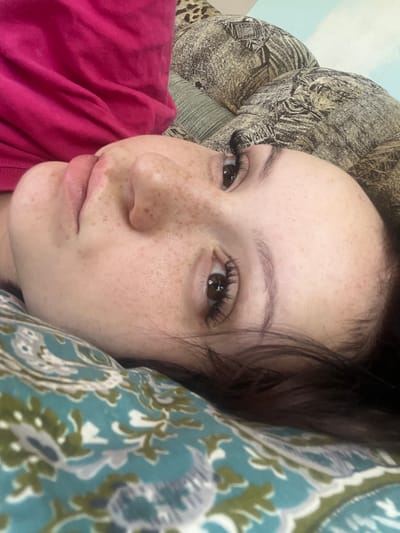 The image is a close-up, portrait-style photograph of a young person lying down, with their head tilted. The subject has fair skin with freckles, brown eyes, and dark eyelashes. Their hair is dark and appears to be resting on a patterned pillow. The subject is wearing a bright pink garment on their upper body. The background consists of a patterned pillow with a design of teal, green, and white floral or paisley motifs, and another pillow or surface with a leopard print pattern, and a light blue area which could be a wall or sky. The overall setting suggests an indoor environment, possibly a bedroom. There is no discernible information in the image that could indicate the location in the city of Zudilovo, Russia.
