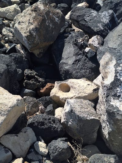 The image shows a pile of rocks of various sizes, shapes, and colors. The rocks are mostly gray and black, with some lighter-colored stones mixed in. One of the lighter-colored rocks has a distinct hole in it. The scene appears to be located in a natural environment, such as a rocky hillside or a riverbed. There are no people or animals visible in the image. The image was captured in Aniavan, Armenia.