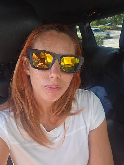 A person with red hair and freckles is wearing sunglasses with yellow lenses and a black frame. The person is wearing a white t-shirt and is sitting in what appears to be a car. The interior of the car is black leather. The reflection in the sunglasses shows a building and some greenery. The person is located in the city of Key Largo, United States of America. Through the car window, trees and cars are visible.