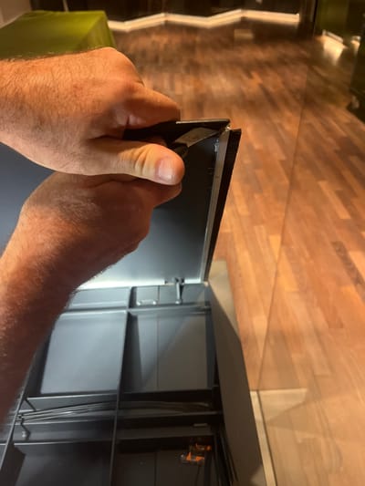 This is a close-up, vertically oriented photograph showing a person's hands working on a black electronic device with a brush-like strip along its edge. The hands are hairy and appear to be those of an adult male. One hand, on the left, is holding a small blade, possibly a utility knife or scalpel, and is carefully slicing through the brush strip. The other hand is positioned on the device, holding it steady. The background consists of a wooden surface, likely a floor or countertop, with a wood grain pattern. Reflections of light are visible on this surface, suggesting artificial lighting. To the left of the frame, a green cloth-covered object is partially visible. The device itself appears to be a component of a larger appliance, possibly a computer or audio-visual equipment, given the grey internal structure with visible wiring and clips. The overall impression is one of a repair or modification task being performed with precision. No text is visible in the image. The setting appears to be indoors, in a domestic or workshop environment. The lighting suggests it could be daytime or evening. No specific details about weather or strong emotions are discernible. The location context of Cologny, Switzerland, does not provide specific visual cues within the image itself.