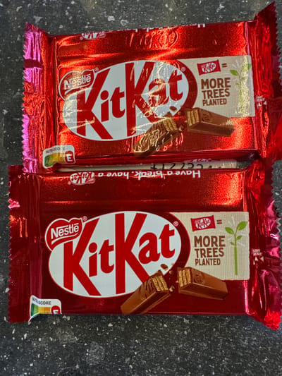 This media file shows two red wrappers for Kit Kat candy bars placed on a dark, speckled surface. The wrappers are shiny and metallic, with the iconic white "Kit Kat" logo prominent on each. The bar on the left is slightly smaller and shows a "More Trees Planted" message with a small plant graphic and a red Kit Kat logo. The bar on the right is larger and also displays the "More Trees Planted" message. Both bars have a small Nestlé logo. In the background, some text is visible on the left bar that reads "Have a break, have a Kit Kat". The lighting suggests an indoor setting, possibly a countertop. The overall mood is neutral, focusing on the product. No people are visible. The location context of Mikhailovka, Russia, is not directly evident from the image itself.
