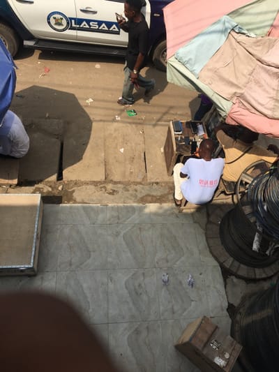 An overhead, high-angle image captures an outdoor urban commercial street scene in Ojo, Lagos State, Nigeria, illuminated by strong sunlight casting sharp shadows.
In the foreground, a light-colored tiled pavement features two light-grey pigeons and two wooden boxes: a large rectangular one with metal trim on the left, and a smaller one with metal clasps on the lower right. Adjacent to this tiled area, a rough concrete strip contains two parallel open drainage gutters. Beyond this, an unpaved, dusty road surface is visible, littered with various debris including paper and a small green circuit board.
On the road, a white pickup truck, identified by "LAGOS STATE SIGNAGE AND ADVERTISEMENT AGENCY" and "LASAA" logos on its side, is partially visible. A Black male, wearing a dark t-shirt, light-colored jeans, and sandals, walks rightward next to the truck while holding a smartphone to his ear.
To the right of the road, two Black males are seated beneath a large, multi-paneled umbrella (pink, light blue, beige fabric sections). The male in the foreground wears a white t-shirt with Chinese characters ("思法教育" - "Sifa Education" / "Rule of Law Education") and "OJO CAMPUS" printed on the back, and light-colored trousers, seated on a low stool facing away from the camera. He is positioned near a wooden stall displaying multiple smartphones. Next to him, a second male in a tan shirt is partially visible, also seated. On the far right, several large spools of black cable are stacked vertically. A dark blue umbrella is partially visible on the far left, shading a person in white clothing.