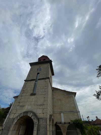A low-angle shot captures a tall, light-stone building, likely a church or bell tower, set against a cloudy sky. The building features an archway, a small window, and a decorative emblem. The tower has a red-tiled roof. The structure is probably located in the city of Tsar Kaloyan, Bulgaria. Tree branches are visible at the top of the frame. The sky is overcast with gray clouds.
