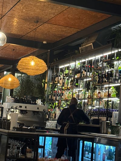 The photograph shows the interior of a restaurant or bar. The wall is clad in brown particleboard, while two decorative light fixtures hang from the ceiling. A man wearing a dark uniform and an apron is standing behind the bar. Behind him, a large wine rack is filled with wine bottles and adorned with greenery. A chilled display cabinet containing beverages and other items sits next to the bar. The photograph was taken indoors, but further information to confirm the location in Paarl, South Africa is not provided.