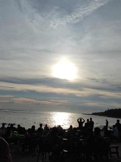 The photo captures a bustling beach scene in Monikeun, Indonesia, bathed in the bright glow of the sun. A group of people are gathered near the shore, some standing and some seated at tables on the sand. Silhouettes of people can be seen swimming or wading in the water. The sky is overcast with textured clouds. A line of trees stretches along the right edge of the frame.